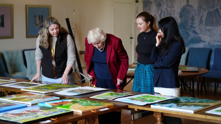 4 judges looking at the shortlisted paintings from the Inspired by Chartwell Painting Competition to determine the winners. The paintings are laid out on tables in the Mulberry room at Chartwell and there is an enlarged reproduction of one of Churchill's paintings in the background.